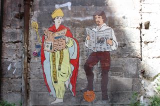Murales "Mission Possible" dell'artista Roxy in the Box che rappresenta San Gennaro e Caravaggio intenti a leggere rispettivamente Il Sole 24 Ore e il New York Times. 